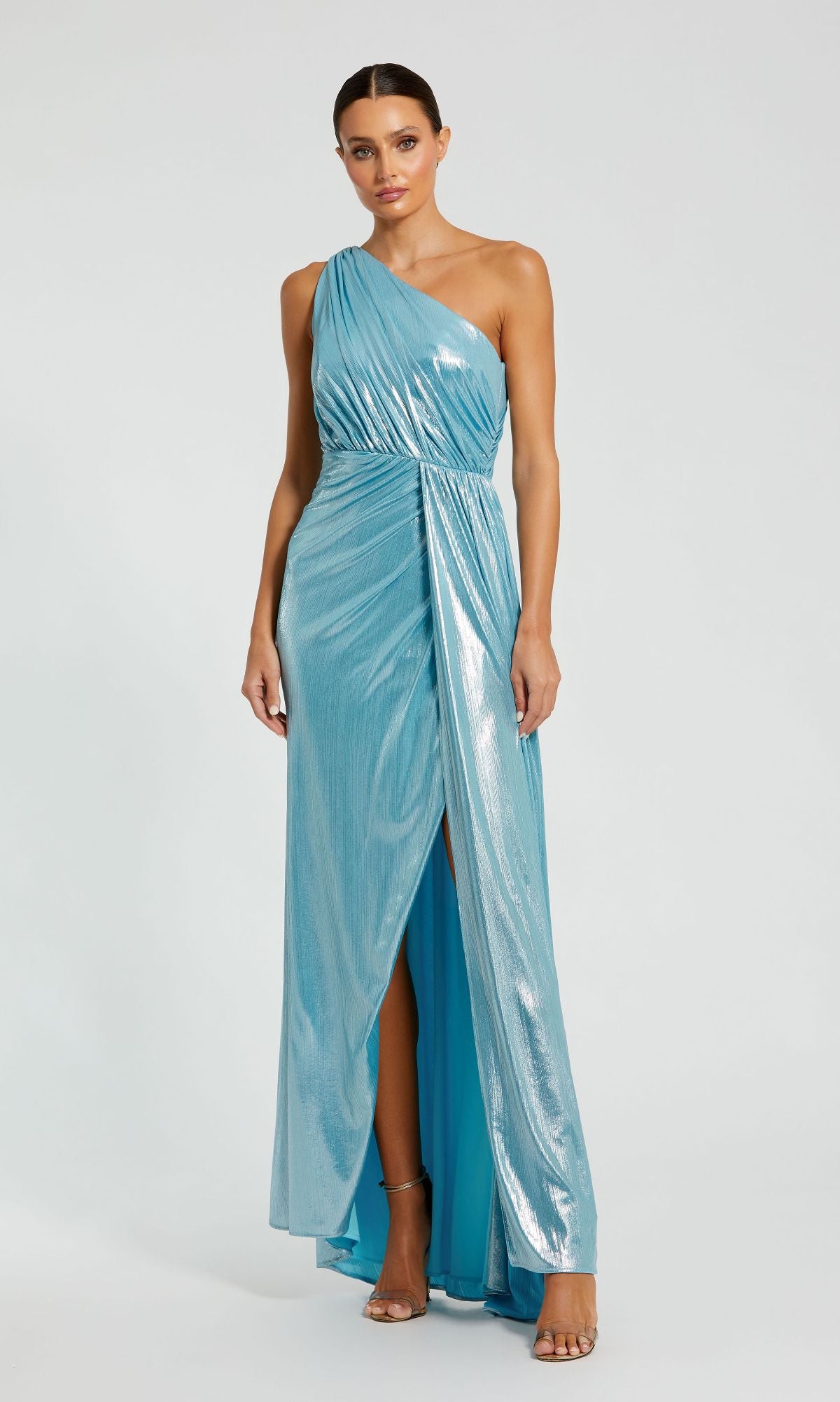 A woman poses against a plain background wearing the Mac Duggal 26537—an elegant one-shoulder metallic formal dress in light blue, featuring a thigh-high slit and floor-length silhouette.